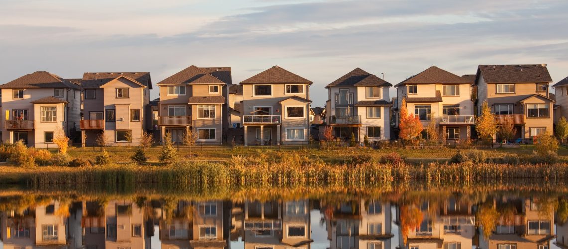 A row of houses reflected in a pond. Horizontal colour image. This classic new neighbourhood or residential region in a large North America City - this is Calgary, Alberta - speaks to the urban sprawl as millions of detached homes cover massive acres or urban area. Here a beautiful wetlands region is close to these homes and provides a nice morning reflection.